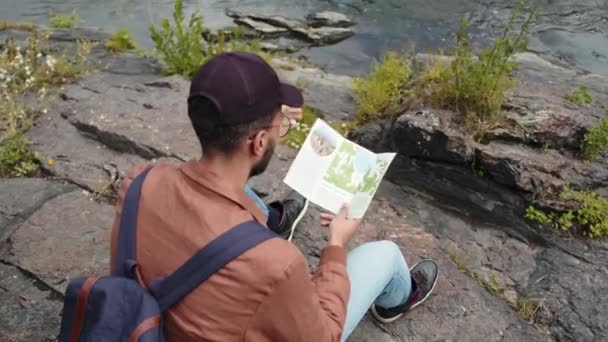 石の上に座っている若い男性Biracial観光客のハイアングルショット パンフレットを読んで周りを見回す — ストック動画