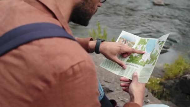 アークショット 若い男性のBiracial観光客が川の近くの石の上に座って昼間の旅行パンフレットを勉強 — ストック動画