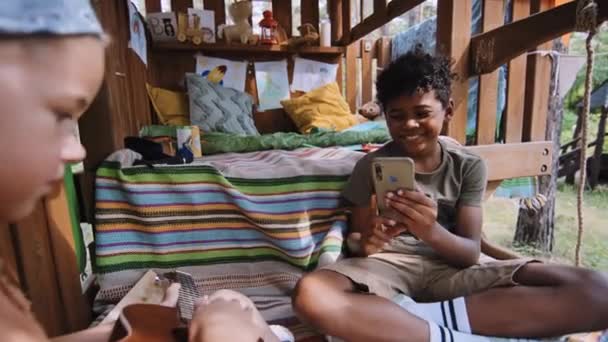 アフリカ系アメリカ人の小さい年齢の男の子のミディアムショット 昼間に木の家でギターを弾く彼のコーカサス人の友人を撮影 — ストック動画
