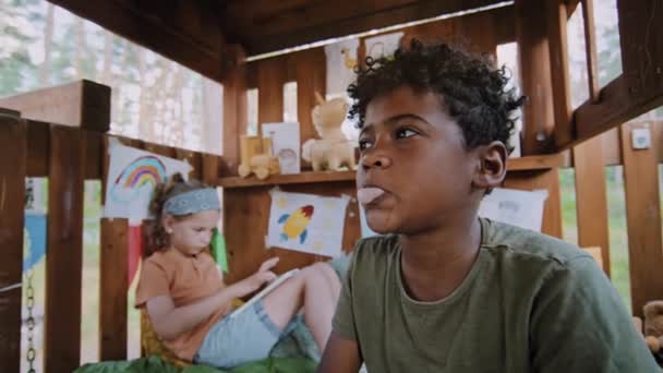 アフリカ系アメリカ人の小さい年齢の少年がガムでバブルを吹き飛ばし 同じ年齢のコーカサス人の少年が木造ハウスの背景でタブレットで遊んでいます — ストック動画