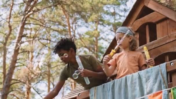 カメラはアフリカ系アメリカ人とコーカサス人の小学生のワイドショットを日中のツリーハウスの上に石鹸バブルで遊びます — ストック動画