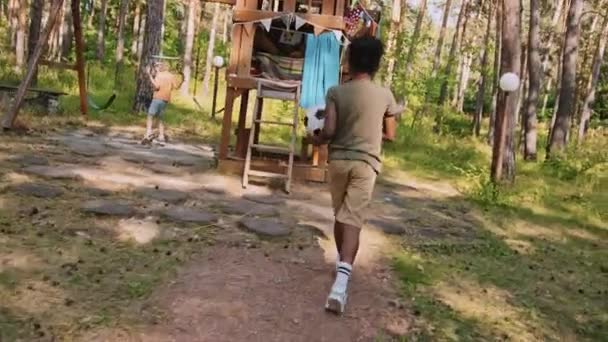 カメラシェイクトラッキングショット アフリカ系アメリカ人とコーカサス人の小さい年齢の男の子は木造ハウスの隣の木の遊び場でサッカーをする — ストック動画
