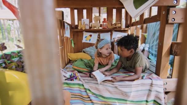 アフリカ系アメリカ人とコーカサス人の小学生のフルショットは 昼間のツリーハウスにフラッシュライト付きの本を読みます — ストック動画
