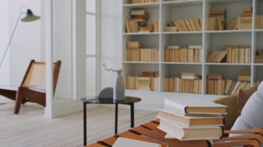 Yüksek tavanlı, minimalist tasarımlı, geniş, çağdaş bir apartman dairesinde tam bir kütüphane fotoğrafı, kanepenin yanında uzanan birkaç kitap, ve kahve masasında kurumuş dallı geometrik vazo.