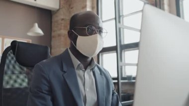 Düşük açılı erkek Afro-Amerikan ofis çalışanı maske takıyor, bilgisayar ekranına bakıyor ve bir şeyler yazıyor.