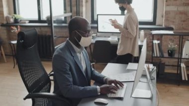 Maske takan ve bilgisayarda çalışan Afrika kökenli Amerikalı bir adamın orta eğim fotoğrafı. Maskeli genç beyaz bir kadın, elinde tabletle ofise gidiyor.