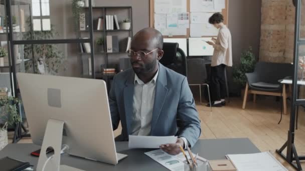 照片中的非洲裔美国男性经理拿着文件在办公桌前工作 而白人女性办公室职员拿着平板电脑在办公室里走着 — 图库视频影像