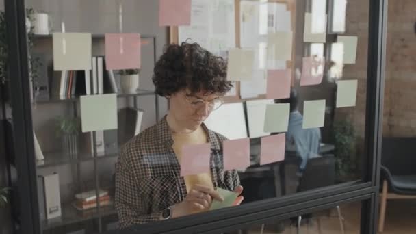 女性の白人マネージャーが昼間のオフィスに透明な壁に粘着性のあるノートを置く中間閉鎖 — ストック動画