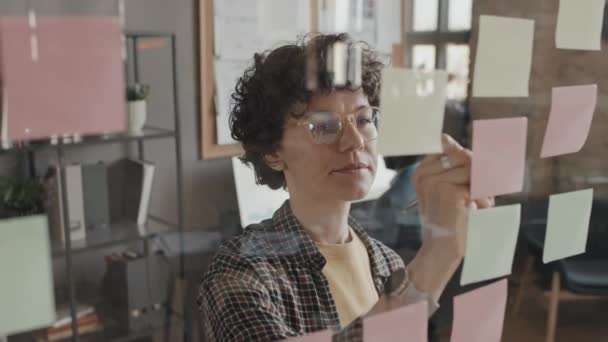 昼間のロフトオフィスに透明な壁に粘着性のあるノートを置く女性のコーカサス事務所の労働者の閉鎖 — ストック動画