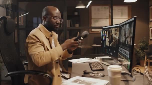 男性アフリカ系アメリカ人編集者のドリー ズームは 夕方にオフィスで机に座り ヘッドフォンを置き 編集プログラムの映像カットで作業する — ストック動画