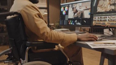Afrika kökenli Amerikalı erkek tekerlekli sandalyeli kullanıcının masa başında oturup klavyeye bir şey yazarken akşam ofiste renk derecelendirme kontrolünü kullanırken görüntüsü.