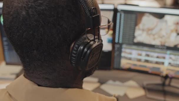 コンピュータに座って編集プログラムで作業するヘッドフォンを調整するアフリカ系アメリカ人の男の頭に焦点を当てる — ストック動画