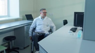 Tekerlekli sandalyedeki erkek bilim adamının soldan görüntüsü laboratuvarda bilgisayarla gündüz vakti çalışma masasına yaklaşıyor.
