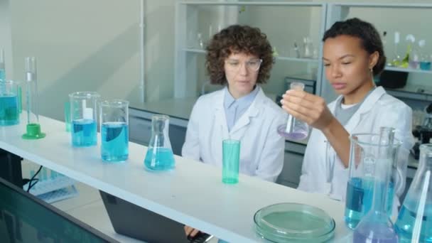 女性生物实验室工作人员从架子上拿出装有紫色液体的瓶子 向白人女性同事展示 — 图库视频影像