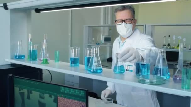 中镜头男性高级科学家戴面具从架子上拿起蓝色液体瓶进行研究 — 图库视频影像