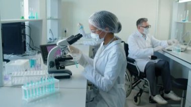 Beyaz laboratuvar önlüklü, gözlüklü ve maskeli bir kadının gündüz vakti laboratuvarda mikroskop altında incelenen araştırmalara bakarken çekilmiş.