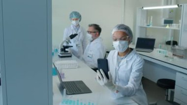 Çift ırklı, maske takan kadın bir laboratuar çalışanı elinde akıllı bir telefonla laboratuvarda video görüşmesi yaparken, arka planda çalışan iki iş arkadaşı.