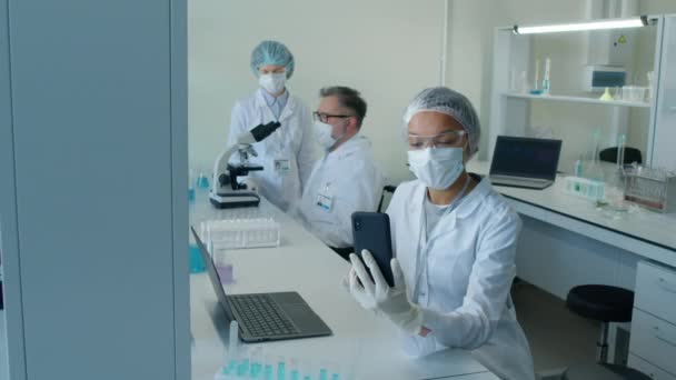 两名在实验室工作的同事 戴着面具 手持智能手机坐在办公桌前 白天在实验室里用视频通话 拍了一张中景照片 — 图库视频影像