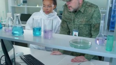 Laboratuvarda durup bilgisayar ekranındaki hücre ve moleküllerin resimlerine bakan asker ve kadın bilim adamının eğik yay görüntüsü.