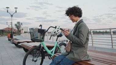 Modern Kafkasyalı iş kadını şık günlük kıyafetler giyiyor sabahın erken saatlerinde bankta oturuyor ve akıllı telefondan bir şeyler izliyor.