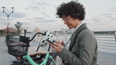 Yetişkin, beyaz, gözlüklü bir kadın sabahın erken saatlerinde bankta oturmuş akıllı telefondan internette bir şeyler arıyor.