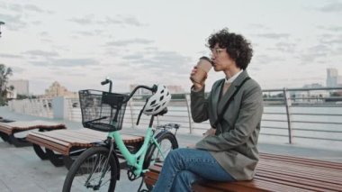 Bayan ofis çalışanı şık günlük kıyafetler giymiş sabahın erken saatlerinde bankta oturmuş kahvesini içiyor.