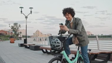 Kafkasyalı bir kadının şık şık, şık kıyafetler giyip bisikletle dışarıda akıllı telefondan internette sörf yaparken orta uzunlukta bir fotoğrafı.