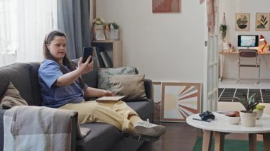 Down sendromlu genç bir kadının kanepede oturup arkadaşlarıyla görüntülü konuşma ve onlara robot resmini gösterme görüntüsü.