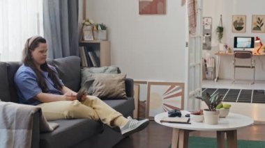 Down sendromlu modern bir kız kanepede oturup dijital tablet kullanarak arkadaşıyla video görüşmesi yapıyor.