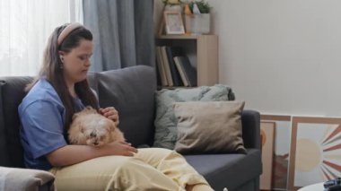 Down sendromlu genç bir kadının oturma odasında kucağında köpekle oturması.