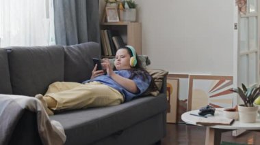 Down sendromlu modern genç kız kulaklık takıyor oturma odasındaki koltukta dinleniyor akıllı telefondan müzik listesi seçiyor.