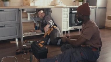 Genç Afrikalı Amerikalı tamirci mutfakta oturmuş, lavabo giderlerini tamir eden Kafkasyalı iş arkadaşına aletler veriyor.