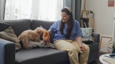 Down sendromlu beyaz bir kadın oturma odasında kanepede oturmuş köpeğiyle oynuyor.