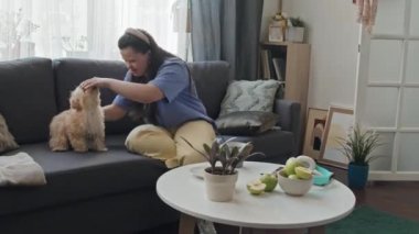 Down sendromlu genç bir kadın oturma odasında oturmuş köpeğiyle eğleniyor.