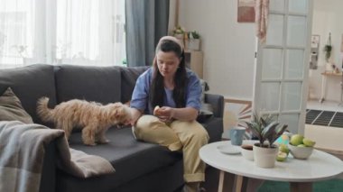 Down sendromlu neşeli genç bir kadın oturma odasında kanepede oturup köpeğine ikramda bulunuyor.
