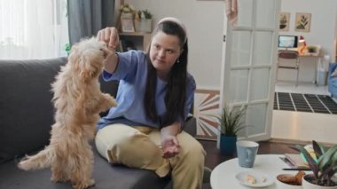 Down sendromlu neşeli genç bir kadın kanepeye oturmuş köpeğine numaralar öğretiyor ve ona hediyeler veriyor.
