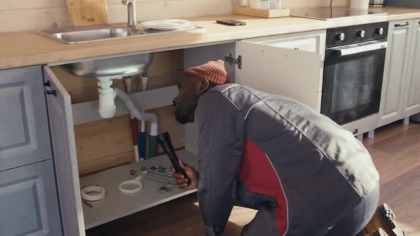 在他成熟的高加索同事与客户讨论工作计划时 非洲裔美国管道工用手电筒发现厨房水池下的管子有问题 — 图库视频影像
