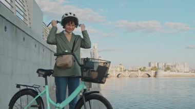 Nehir kıyısında bisikletle dikilen, kaskını çıkarıp kameraya gülümseyen modern beyaz kadının orta boy portresi.
