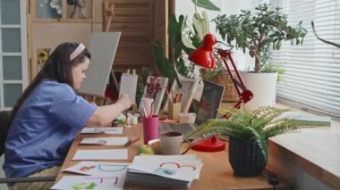 Yan görüş, engelli genç bir kadının masa başında oturup kağıt üzerine bir şeyler boyadığını gösteriyor.