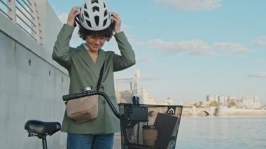 Nehir kenarında bisikletle dikilen modern iş kadınının orta boy portresi. Kameraya gülümseyen kaskını çıkarıyor.