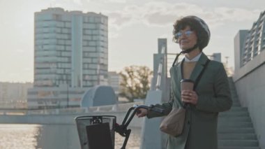 Kafkaslı, bisiklet kaskı takan mutlu beyaz kadın ofis çalışanının orta boy portresi. Sabah nehir kıyısında duran kahve bardağını tutuyor.