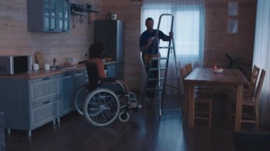 Engelli bir kadına ait evde çalışan iki farklı ırktan işçi var. Biri mutfakta lamba değiştiriyor.