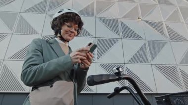 Kıvırcık saçlı, bisiklet kaskı takan beyaz bir kadının akıllı telefondan mesaj attığı düşük açılı bir fotoğraf.