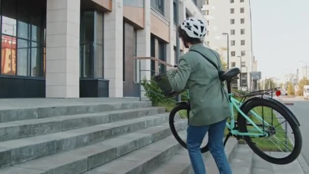 スマートなカジュアルな服とヘルメットを着て 外の階段を歩いているときに自転車を運ぶ認識できない女性のショットに従ってください — ストック動画
