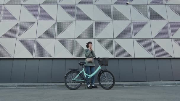 ヘルメットを離れて周りを見回す灰色のオフィスの建物に対して彼女の自転車と立っている長い女性のショット — ストック動画