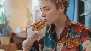 Modern kafede protez kollu sandviç yiyip dizüstü bilgisayarından bir şeyler izleyen, engelli Z jenerasyonundan şık genç bir kız.