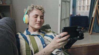 Kısa saçlı, kol protezi takan, oturma odasında dinlenen kulaklıkla müzik dinleyen ve akıllı telefondan internette sörf yapan zarif bir kız.