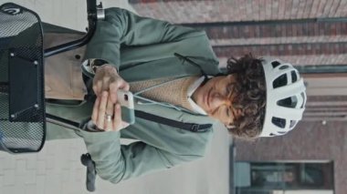 Dikey çekimde, kıvırcık saçlı, şık giyinen, gözlüklü ve kasklı, akıllı telefondaki bisiklet rotasını kontrol eden modern beyaz bir kadın görülüyor.