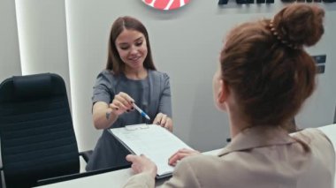 Modern klinikteki kayıt masasında çalışan genç resepsiyonistin hastanın belgeyi nasıl imzalayacağını açıklarken seçici odak noktası.