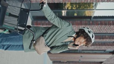 Dikey orta boy beyaz kadın şık günlük kıyafetler giyiyor, gözlük takıyor ve bisiklet kaskı telefonda sohbet ediyor.